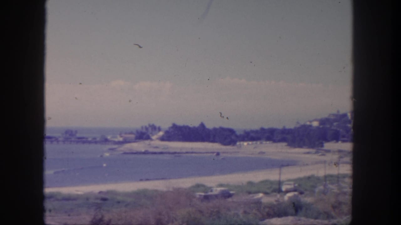 Elevated coastal view with calm water, sandy beach, distant pier, trees, and dark window-frame edges.