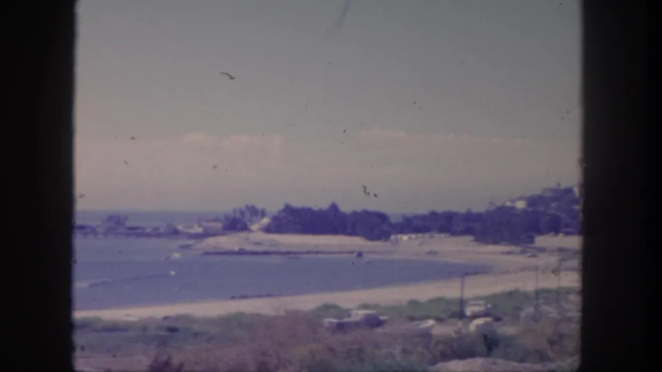 1962 San Pedro coastal home-movie frame showing a beach, calm water, distant pier, parked cars, and dark vertical frame borders.