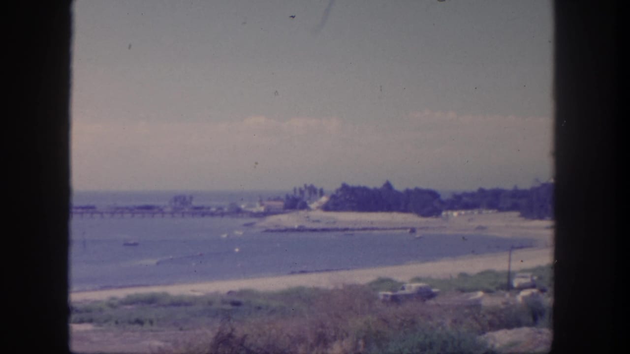 Beachfront and bay view with pier in the distance, shoreline vegetation, parked cars, and window-frame borders.