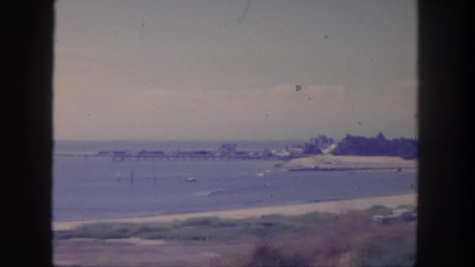 1962 San Pedro home-movie frame showing calm bay water, a narrow beach, distant pier structure, shoreline brush, and soft overcast light.