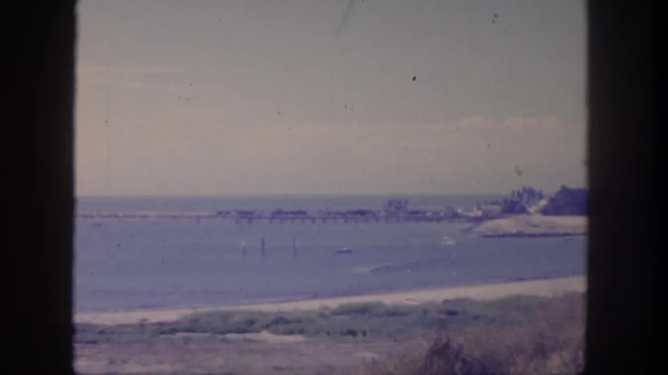 1962 San Pedro coastal view with open water, sandy shoreline, distant pier or jetty, low beach vegetation, trees, and visible 8mm film texture.