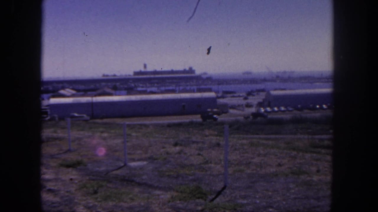 Harbor-side military base view with long buildings, fence posts, parked vehicles, and a distant ship.