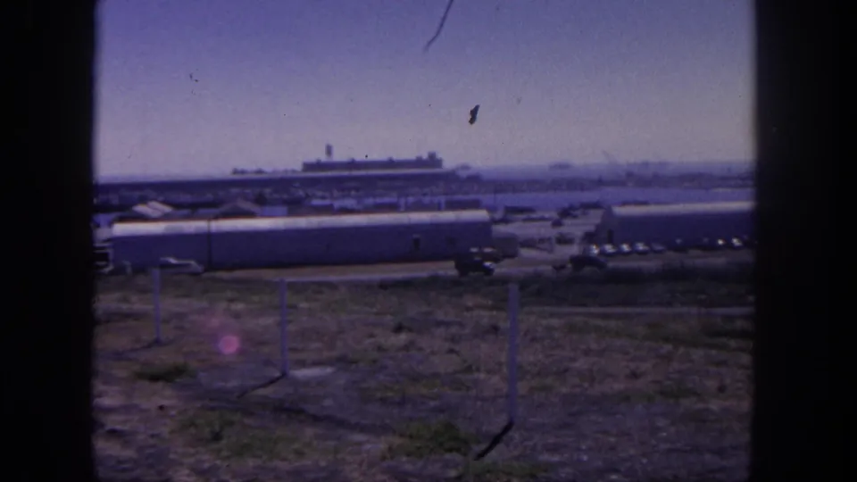 1962 San Pedro base view through dark framing, showing low buildings, fence posts, parked vehicles, dry ground, and a ship in the harbor.