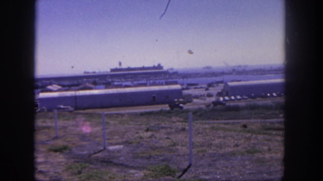 San Pedro, California base landscape with barracks-like buildings, vehicles, fence posts, and a large harbor ship under a pale sky.