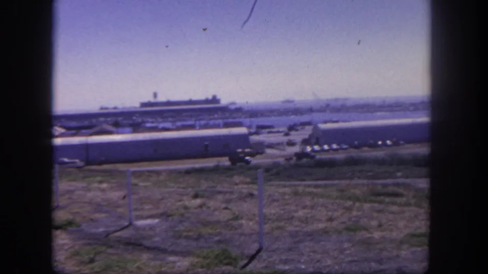 1962 home-movie frame of San Pedro military-base buildings and parked vehicles, with dry foreground and a distant harbor ship.