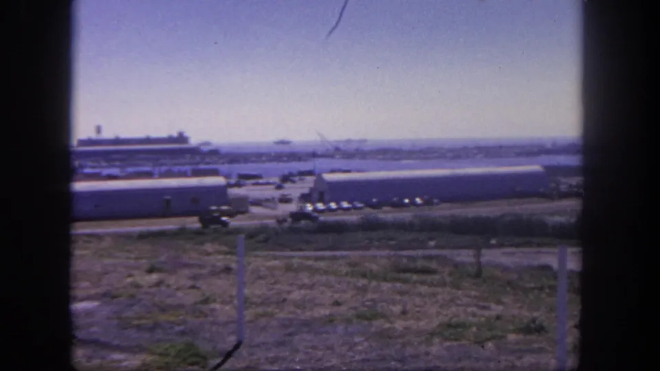 San Pedro base and harbor view from 1962, showing long low structures, parked vehicles, foreground posts, and a large ship beyond.