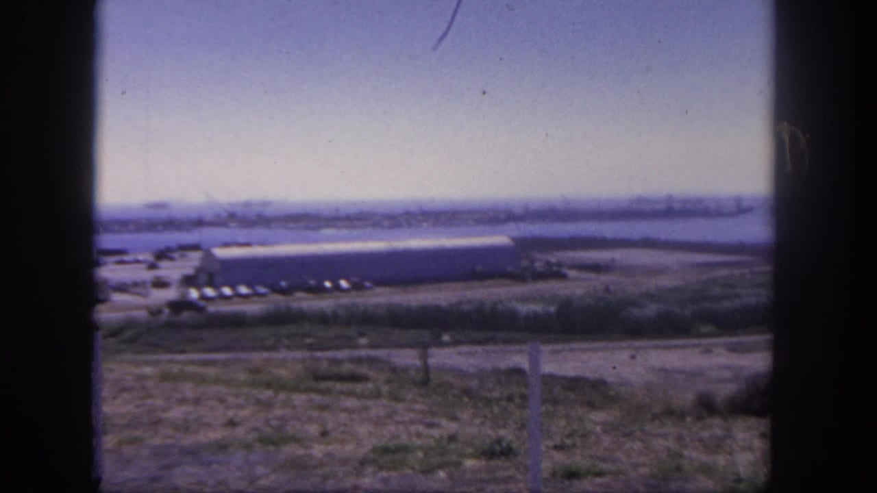 Wide 1962 San Pedro harbor frame showing distant ships and cranes beyond a shoreline building, parked cars, dry grass, and dark film edges.