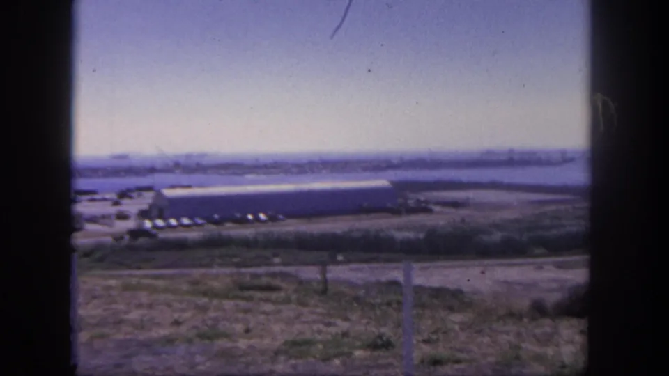 Wide 1962 San Pedro harbor frame showing distant ships and cranes beyond a shoreline building, parked cars, dry grass, and dark film edges.