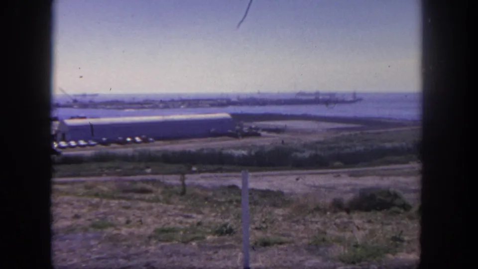 San Pedro harbor shoreline in 1962 with a pale waterfront building, parked cars, water, distant cranes, dry vegetation, and fence posts.