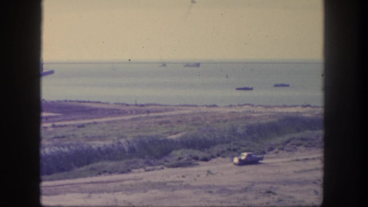 1962 San Pedro home-movie frame showing a car on a coastal road below an elevated viewpoint, with multiple ships visible offshore.