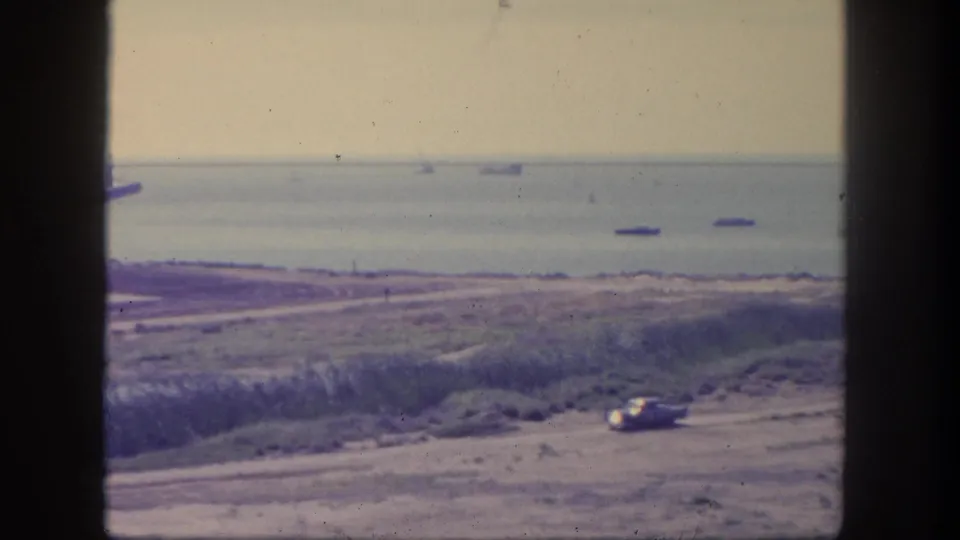 1962 San Pedro home-movie frame showing a car on a coastal road below an elevated viewpoint, with multiple ships visible offshore.