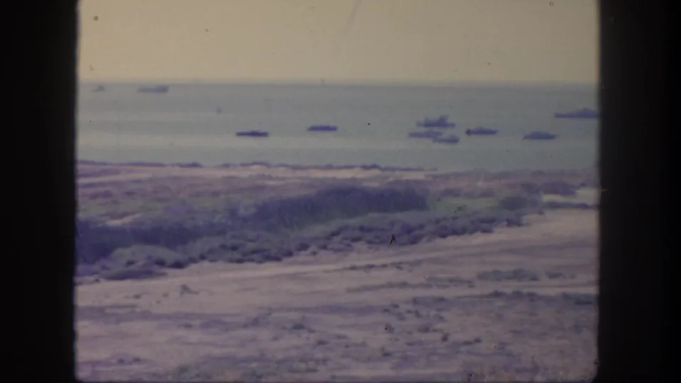 1962 San Pedro 8mm frame showing scrubby coastal ground, a shoreline, and multiple ships offshore under bright daylight.