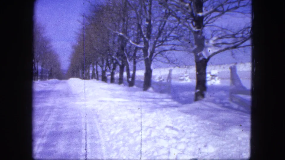 1963 8mm frame from Proctor, Minnesota showing a blue-tinted snowy road bordered by trees and open winter ground.