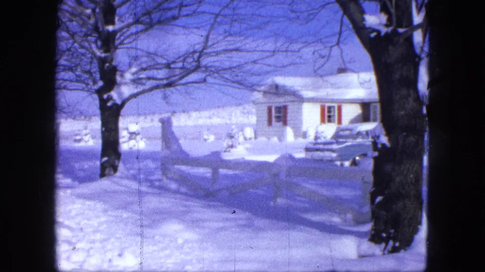 1963 Proctor, Minnesota winter home-movie frame with a white house, red shutters, parked vehicle, fence, and large foreground tree.