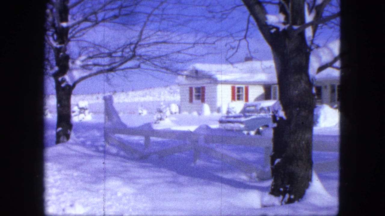 Snow-covered house behind a white fence with a large bare tree and parked car nearby.