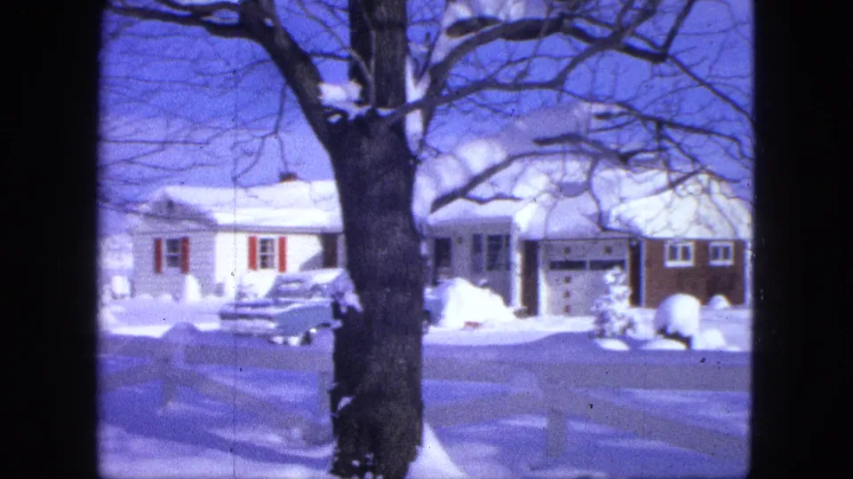 1963 home-movie view of a snow-covered Minnesota house with red shutters, a large foreground tree, and a parked car.