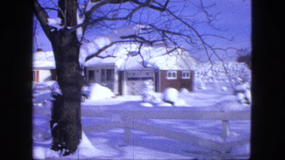 Proctor winter home-movie frame showing a house, garage, large tree, white fence, and deep snow under blue sky.