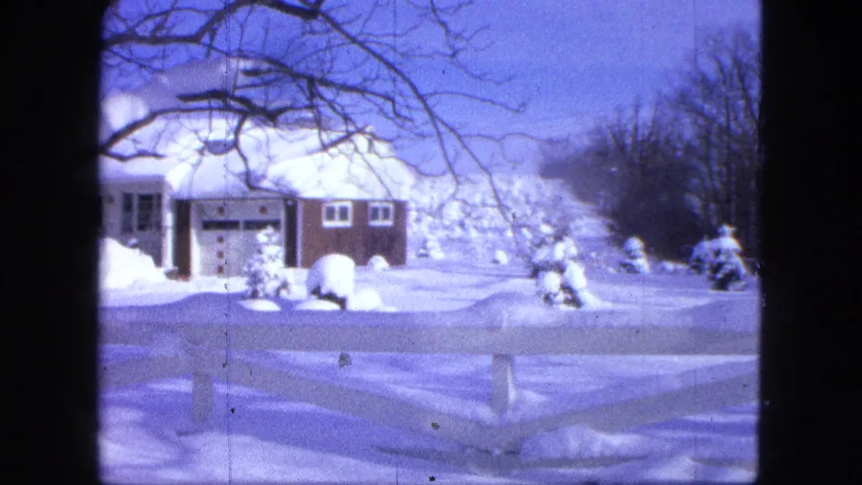 Snow-covered Proctor house in a 1963 8mm frame, with dark vertical edges and bare branches framing the winter view.