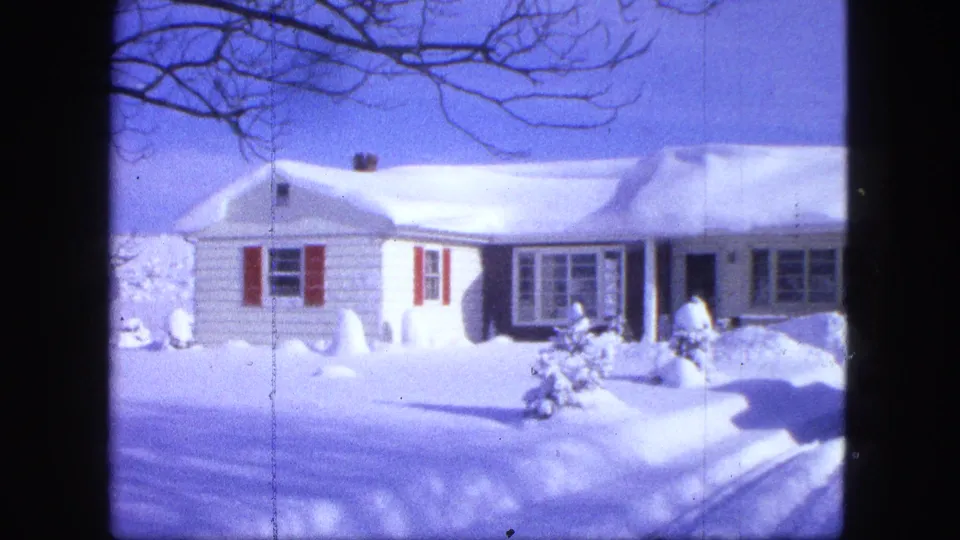 1963 Proctor, Minnesota frame showing the white house facade, red shutters, bare branches, and deep snowbanks around the driveway.