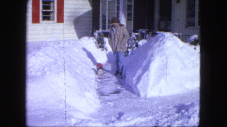 1963 Proctor, Minnesota frame with a woman and small dog on a cleared path between high snowbanks outside a house.