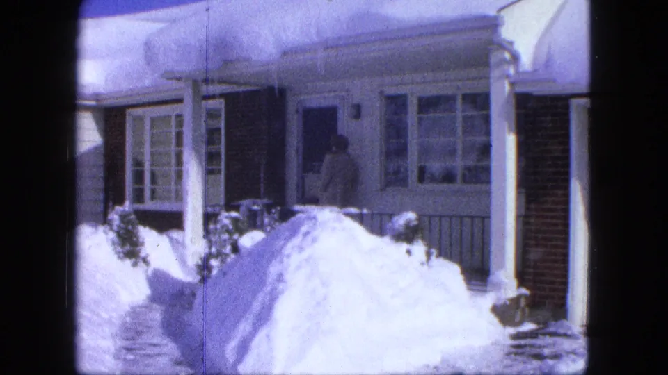 1963 winter home-movie frame from Proctor, Minnesota showing a person entering a front door while snowbanks fill the foreground.