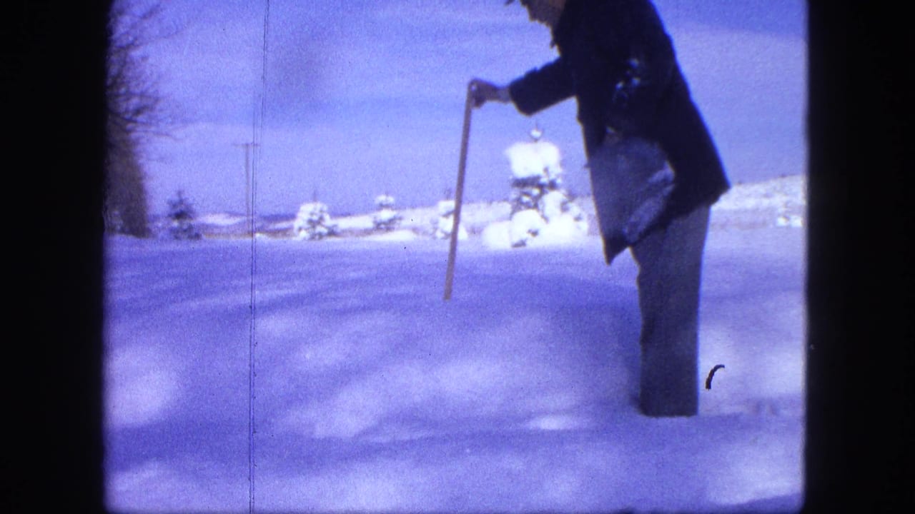A man in a dark coat and hat stands in a snowy Proctor field, holding a long stick toward the snow surface.