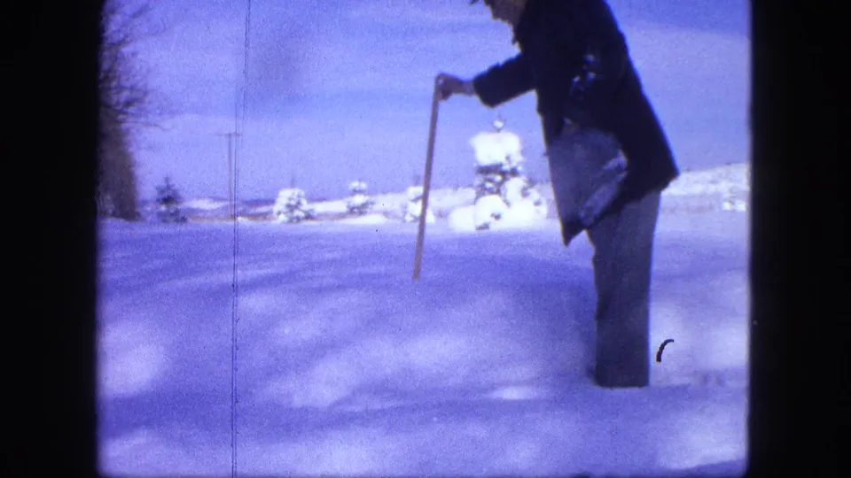 A man in a dark coat and hat stands in a snowy Proctor field, holding a long stick toward the snow surface.