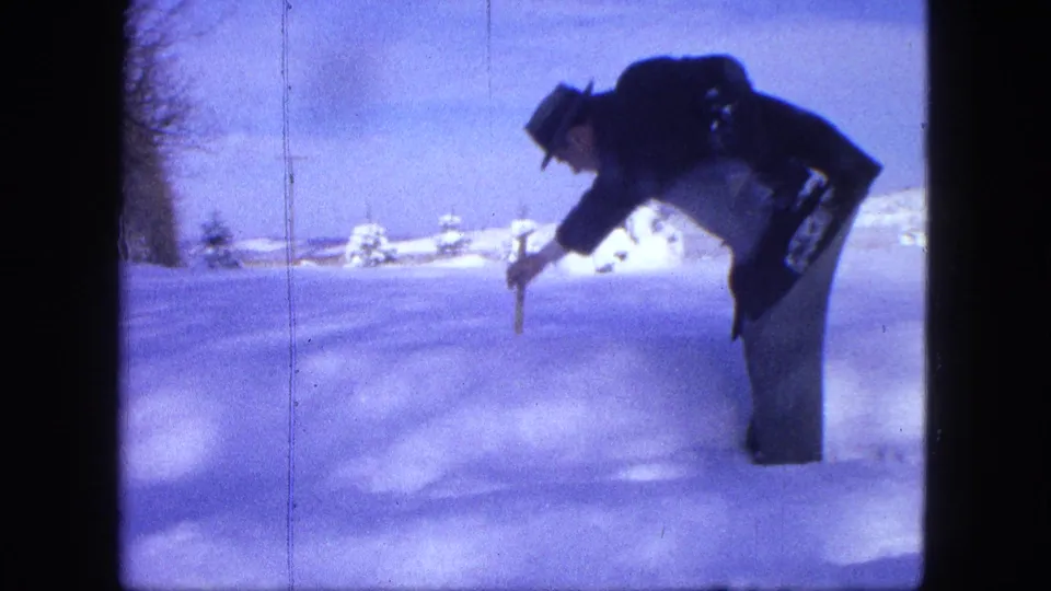 A man in winter clothing leans over a stick set into the snow, with snow-covered shrubs and a utility pole in the background.