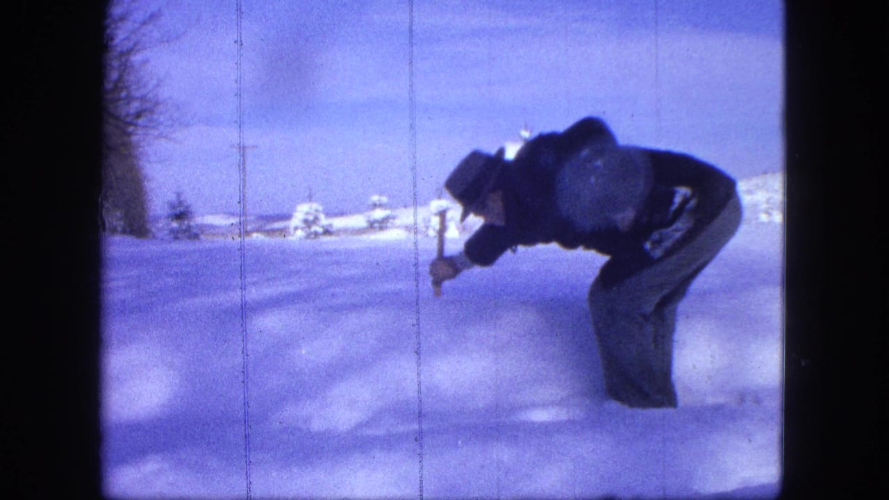 Man bends over deep snow with a stick in a snowy rural landscape.