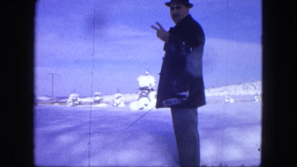 A man in 1963 Proctor winter footage faces the camera and raises two fingers while standing in the snow with a pole.