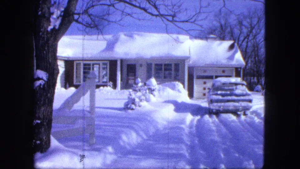 1963 8mm frame in Proctor, Minnesota, showing a snowy residential exterior with garage, buried car, and winter trees.
