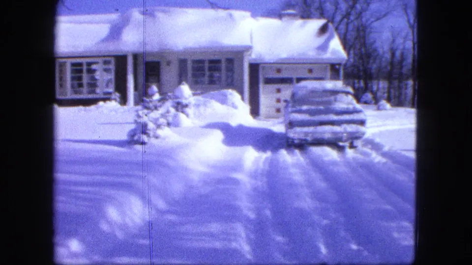 The snowy residential exterior remains framed on the house, garage, driveway tracks, and car under heavy winter snow.