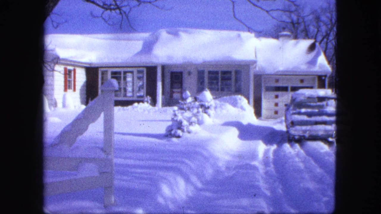Snow-covered white house with red shutters, fence post, driveway, and parked car.