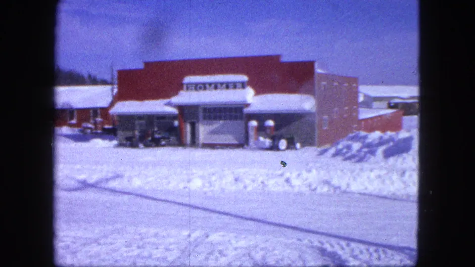 1963 Proctor, Minnesota home-movie view of the red HOMMER storefront framed by deep snow and parked vehicles.