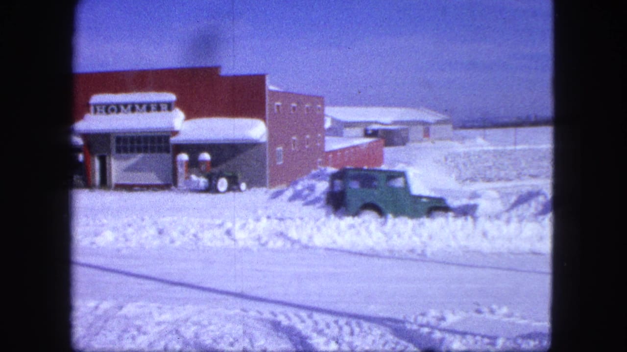 A green vehicle drives through snow near the red brick HOMMER building in this 1963 Proctor, Minnesota home-movie frame.