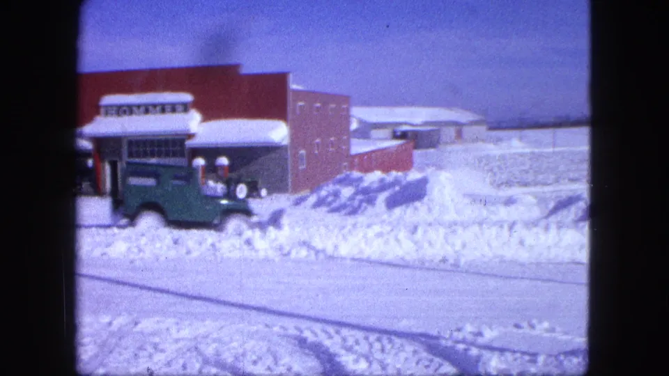 The green vehicle moves across the snow-covered roadside while the Proctor brick building and HOMMER sign remain partly visible.