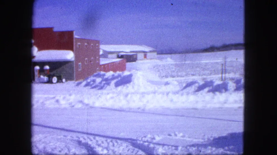 A quiet 1963 Proctor, Minnesota winter roadside view remains after the passing vehicle exits, with the brick building and snowy open ground visible.