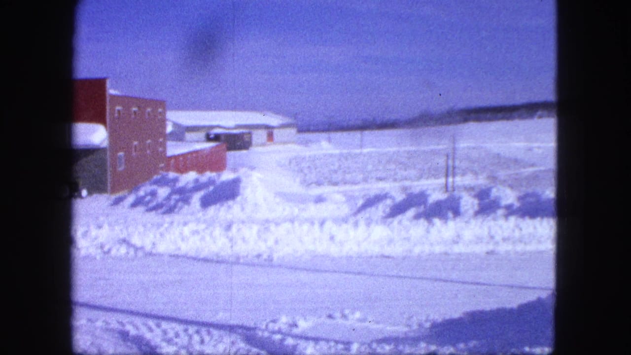 Snowy field with a red brick building, snowbanks, utility poles, and blue sky.