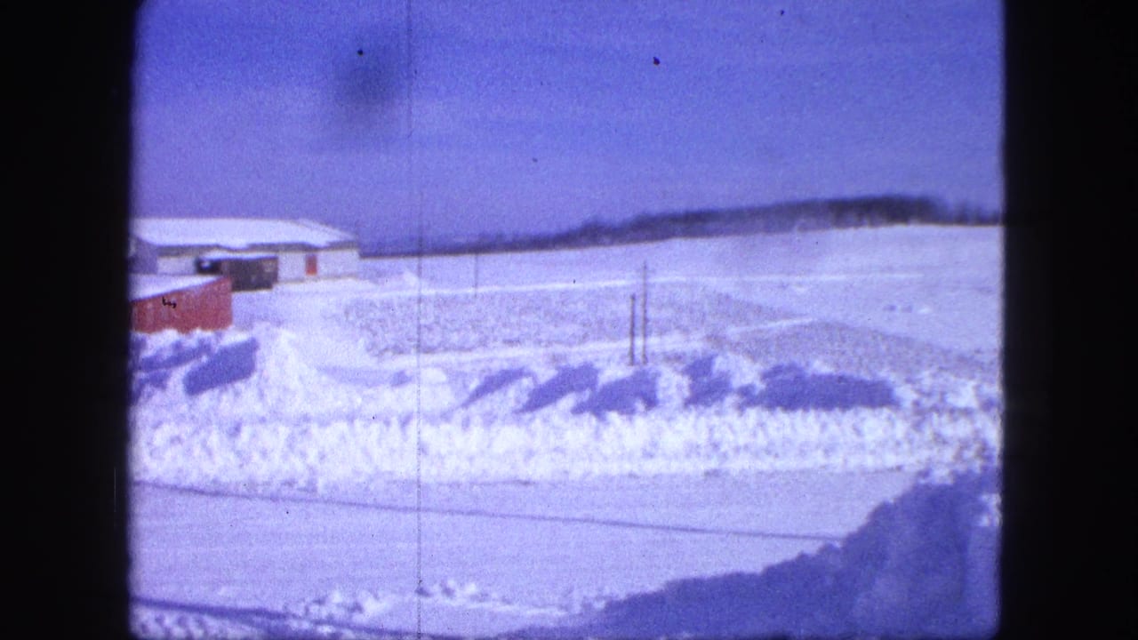 1963 Proctor, Minnesota frame with open snow-covered ground, low buildings, and a roadside snowbank under a blue sky.