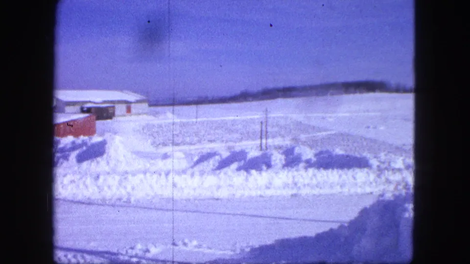 1963 Proctor, Minnesota frame with open snow-covered ground, low buildings, and a roadside snowbank under a blue sky.