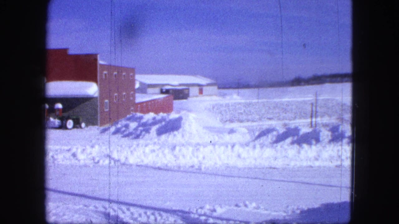 Snowy road facing a red brick storefront with snowbanks and a plow vehicle partly visible at left.