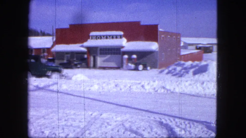 1963 Proctor, Minnesota frame showing a green plow Jeep passing the red brick HOMMER storefront amid heavy snowbanks.