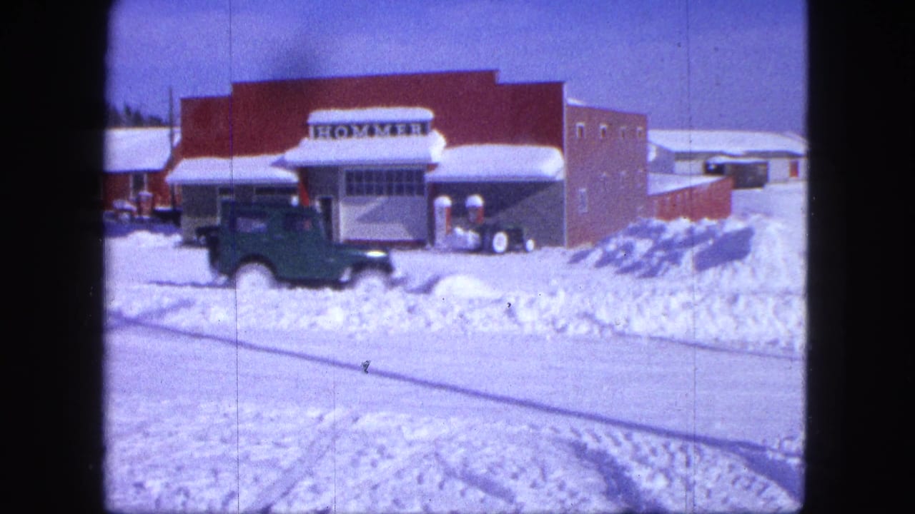 Green car on a snow-covered road near a red building with a HOMMER sign.