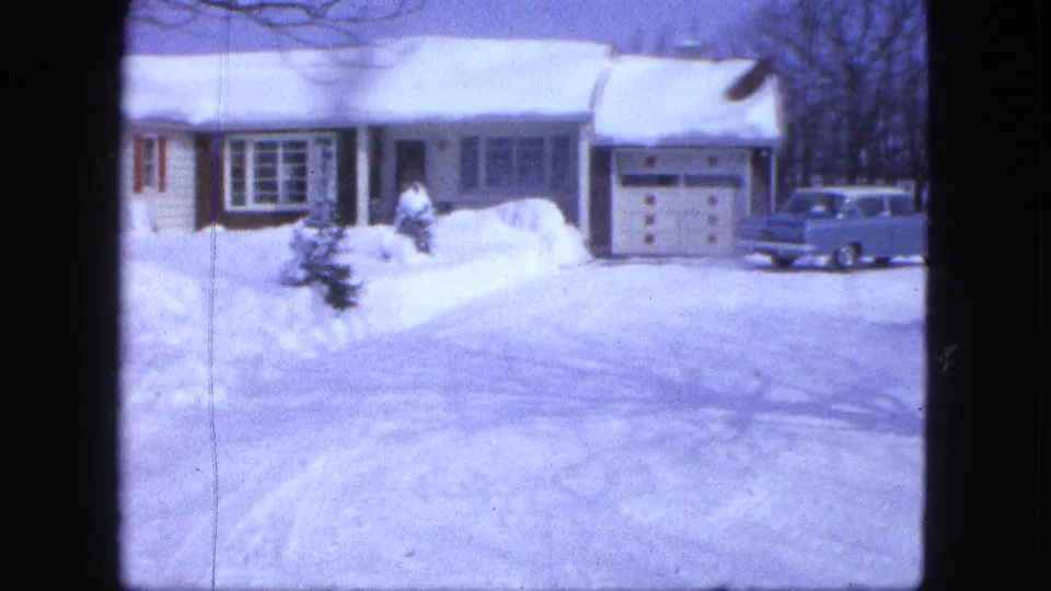 1963 Proctor, Minnesota home-movie frame showing a blue mid-century car near an attached garage, with deep snow around the driveway.