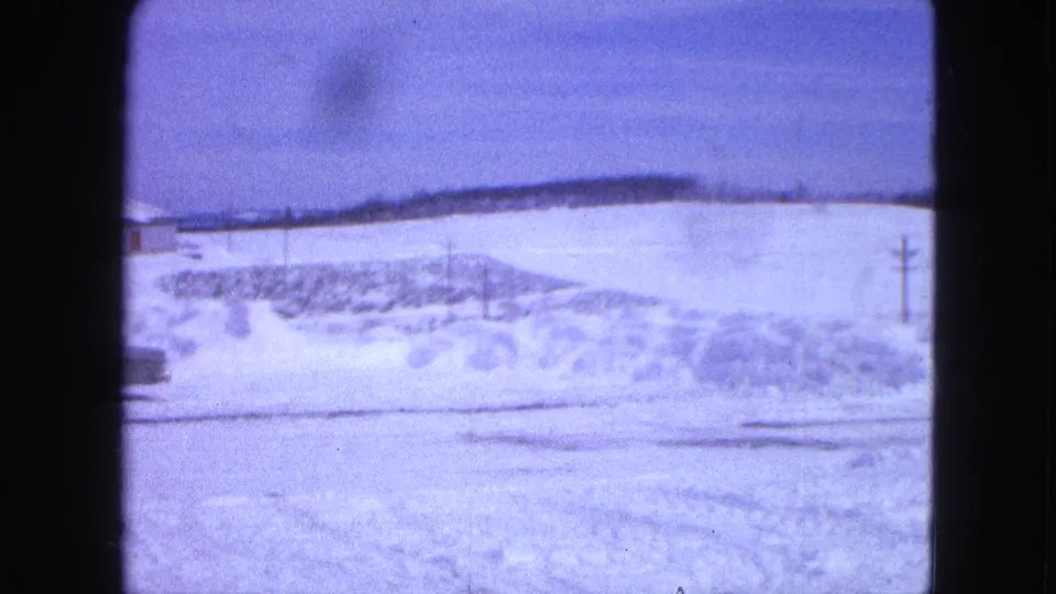 1963 Proctor, Minnesota frame looking past roadside snowbanks toward open snow-covered fields and a distant tree line.
