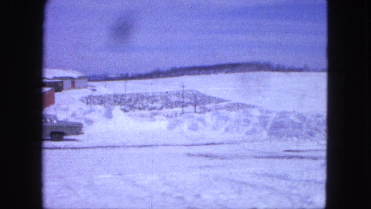 Snowy roadside with a partial car at left and red building edge beyond deep snowbanks.