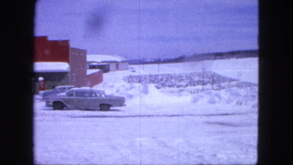Silver classic sedan passes a red commercial building in snowy 1963 Proctor, Minnesota surroundings.