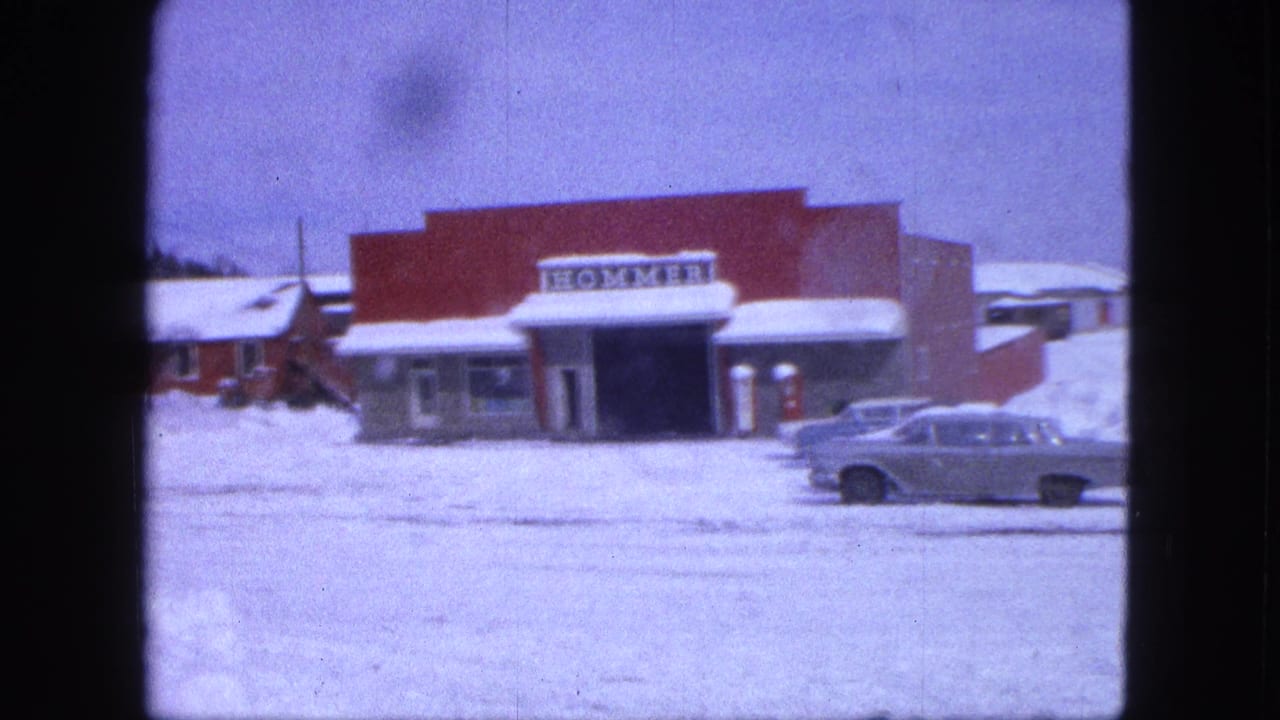 Snowy street facing a red storefront with a HOMMER sign and a parked 1960s car.