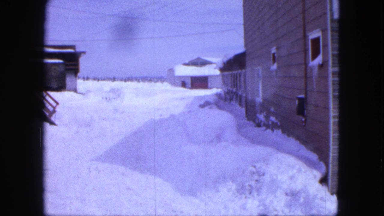 1963 Proctor, Minnesota home-movie frame showing a deep snowdrift beside wooden buildings and a small outbuilding under utility lines.