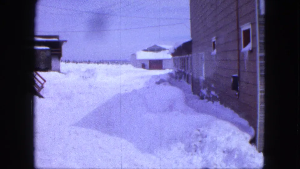 1963 Proctor, Minnesota home-movie frame showing a deep snowdrift beside wooden buildings and a small outbuilding under utility lines.